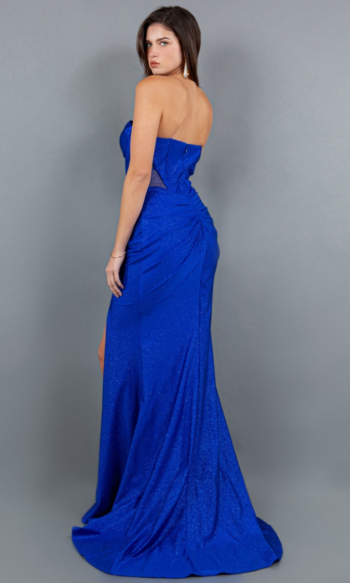 A woman poses with her back mostly to the camera in a strapless, floor-length royal blue Long Glitter Prom Dress (Cinderella Couture 8094J) featuring a corset-style bodice, set against a plain gray background.